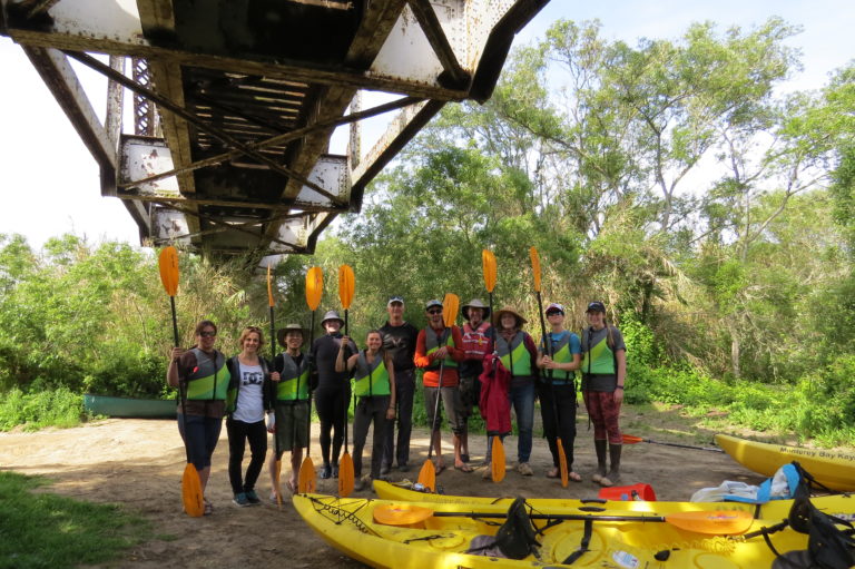 A dozen people stand under a bridge by a river with paddles in their hands and yellow kayaks lay in front of them.