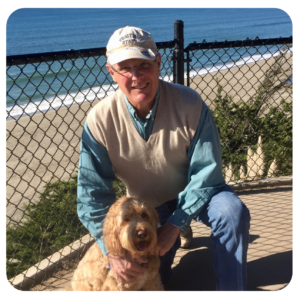 A man crouches down to smile beside his curly small dog with a fence in the background and sand and the ocean in the distance.