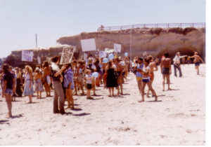 Dozens of people in the early 1980's gather for a protect on a beach with signs and blue balloons and a bluff in the background.