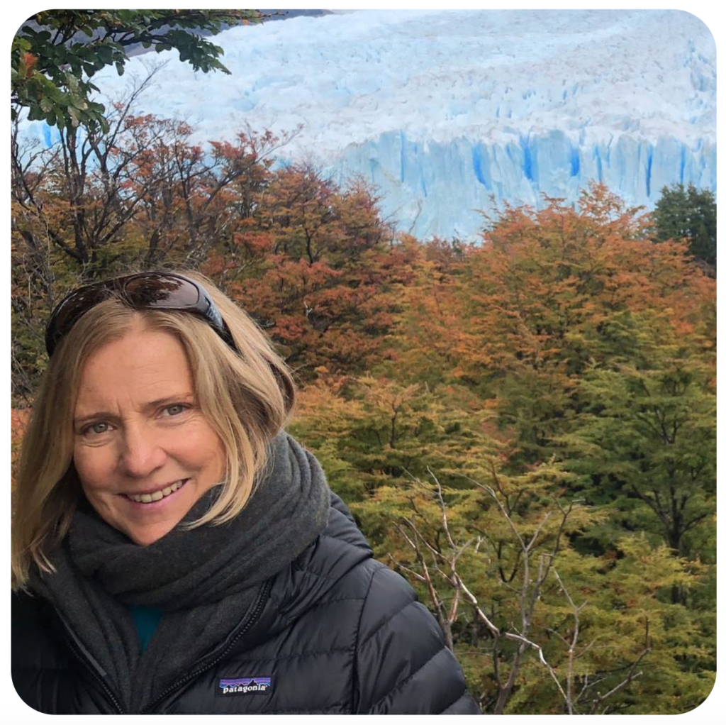 A smiling blond woman in a black down coat stands before green and orange trees with glaciers in the far distance.