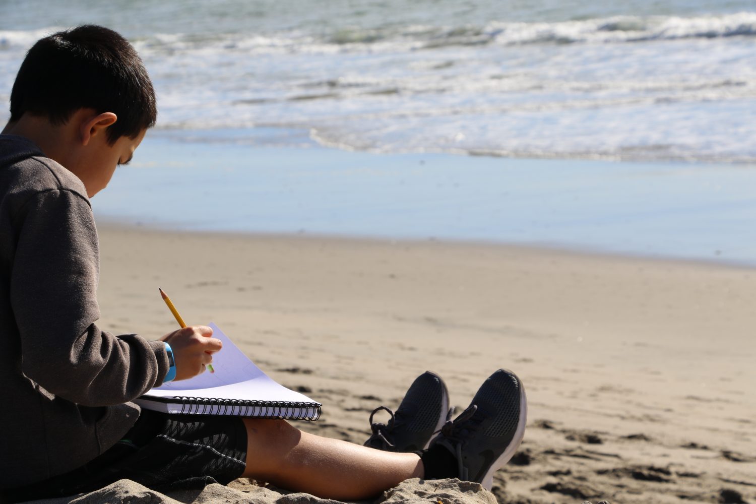 An elementary boy sits on Twin Lake State Beach near the water and writes in pencil in a journal.