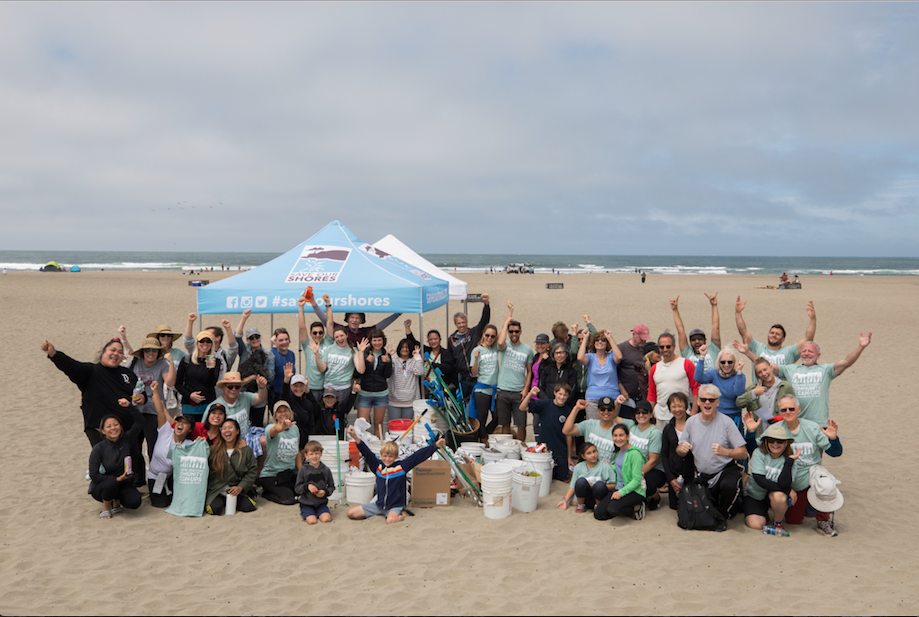 A couple dozen Alain Pinel Realtor team members throw their hands up in celebration on the beach after a beach cleanup.